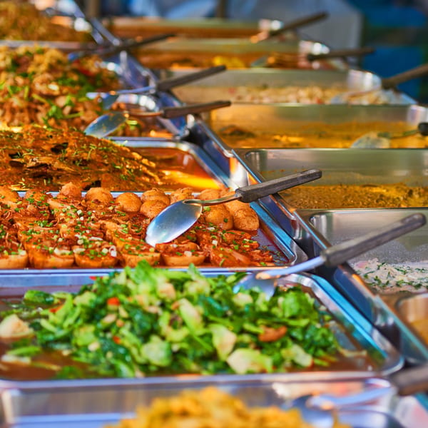 A buffet line with various hot dishes, including meat, vegetables, and sauces, in metal serving trays.